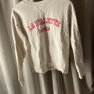 Vit sweatshirt med tryck - Säljer en vit sweatshirt med rött tryck 'La Villette Paris' på bröstet. Tröjan har långa ärmar och en rund halsringning. Perfekt för en avslappnad stil.
