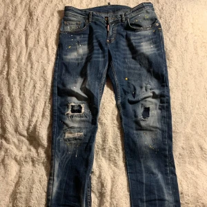 Blå jeans från DSQUARED2 - Snygga blå jeans från DSQUARED2 med slitna detaljer och färgstänk för en snygg design. De har en klassisk femficksdesign och en läderpatch med logotyp baktill. Perfekta för en avslappnad stil.