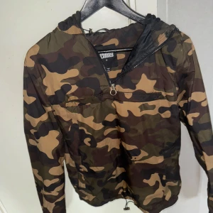 Camouflage vindjacka med huva - Säljer en camouflage vindjacka från Classics i storlek S. Jackan har en praktisk huva och dragkedja framtill. Perfekt för utomhusaktiviteter och har en justerbar nederkant för bättre passform.