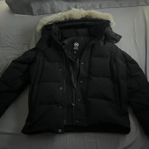 Canada Goose Wyndham - Säljer en svart dunjacka från Canada Goose med pälskantad huva. Jackan har en klassisk design med dragkedja och knappar framtill. Perfekt för kalla vinterdagar. Använd 5-10 gånger går att ta av och på pälsen med en zipper. den har inga defekter ny pris 10.000kr mitt pris 3500kr. Storlek S passar mig perfekt är 180 väger 60kg. Pris går att diskuteras