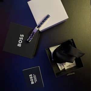 Svart och silver armband från Boss - Elegant armband från Boss i svart och silver. Armbandet har en stilren design med länkar och Boss-logotypen ingraverad på en av länkarna. Perfekt för att ge en sofistikerad touch till din stil. Säljer armbandet för att den är för stor för mig och har även med kvittot.