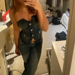 Jeanskorsett - Snygg jeanskorsett med knappar framtill. Den har en tight passform och är perfekt för att ge en cool och trendig look. Korsetten är ärmlös och har en klassisk denimfärg.