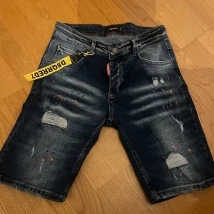 Blå jeansshorts från Dsquared2 - Snygga blå jeansshorts från Dsquared2 med slitna detaljer och en cool, avslappnad look. Shortsen har en klassisk femficksdesign och en märkestagg baktill. Perfekta för en trendig stil!