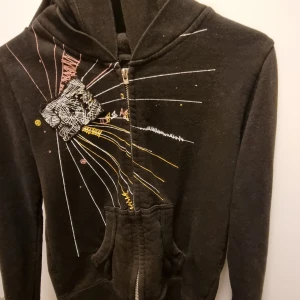 Svart hoodie med färgglada detaljer - Säljer en svart hoodie med dragkedja och unika färgglada broderade detaljer på framsidan. Tröjan har en bekväm passform och en praktisk ficka framtill. Perfekt för en avslappnad stil.
