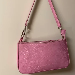 Rosa axelväska med krokodilmönster - Snygg rosa axelväska i syntet med krokodilmönster. Väskan har en dragkedja upptill och en extra dragkedja på framsidan för småsaker. Perfekt för att ge din outfit en färgklick!