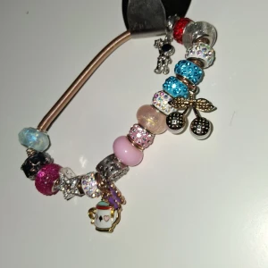 Färgglatt armband med berlocker - Charmigt armband med flera färgglada pärlor och berlocker, inklusive körsbär och en julkopp. Pärlorna är i olika färger som blå, rosa och silver, och vissa är glittriga. Perfekt för att ge en personlig touch till din stil.