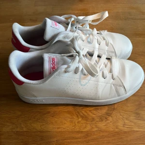 Vita sneakers från Adidas - Snygga vita sneakers från Adidas med rosa detaljer på hälen och logotypen. Skorna har klassisk snörning och en stilren design som passar perfekt till vardags. De är tillverkade i syntetmaterial och har en bekväm passform. Storlek 39 1/3 passar mig som oftast har 39/40 beroende på märke 