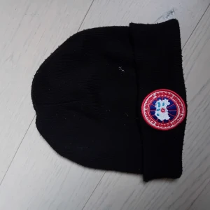 Svart mössa från Canada Goose - Snygg svart mössa från Canada Goose med ett broderat märke framtill. Perfekt för att hålla värmen under kalla dagar. Ej säker på att den är äkta därav de låga priset. Helt okej skick, behöver ba tvättas, priset kan diskuteras vid snabb affär
