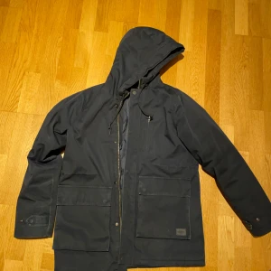 Mörkblå parkajacka med huva från Suit - Säljer en stilren mörkblå parkajacka med huva. Jackan har dragkedja och knappar framtill samt flera praktiska fickor. Perfekt för kyligare dagar med sin långa ärm och skyddande design. Jag är 182 och väger 80kg 