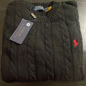 Svart kabelstickad tröja från Polo Ralph Lauren - ”Går ner i pris vid snabb o smidig affär”.  Säljer en stilren svart kabelstickad tröja från Polo Ralph Lauren med det klassiska röda logotypbroderiet på bröstet. Tröjan har en rund halsringning och är perfekt för en elegant och avslappnad look. 