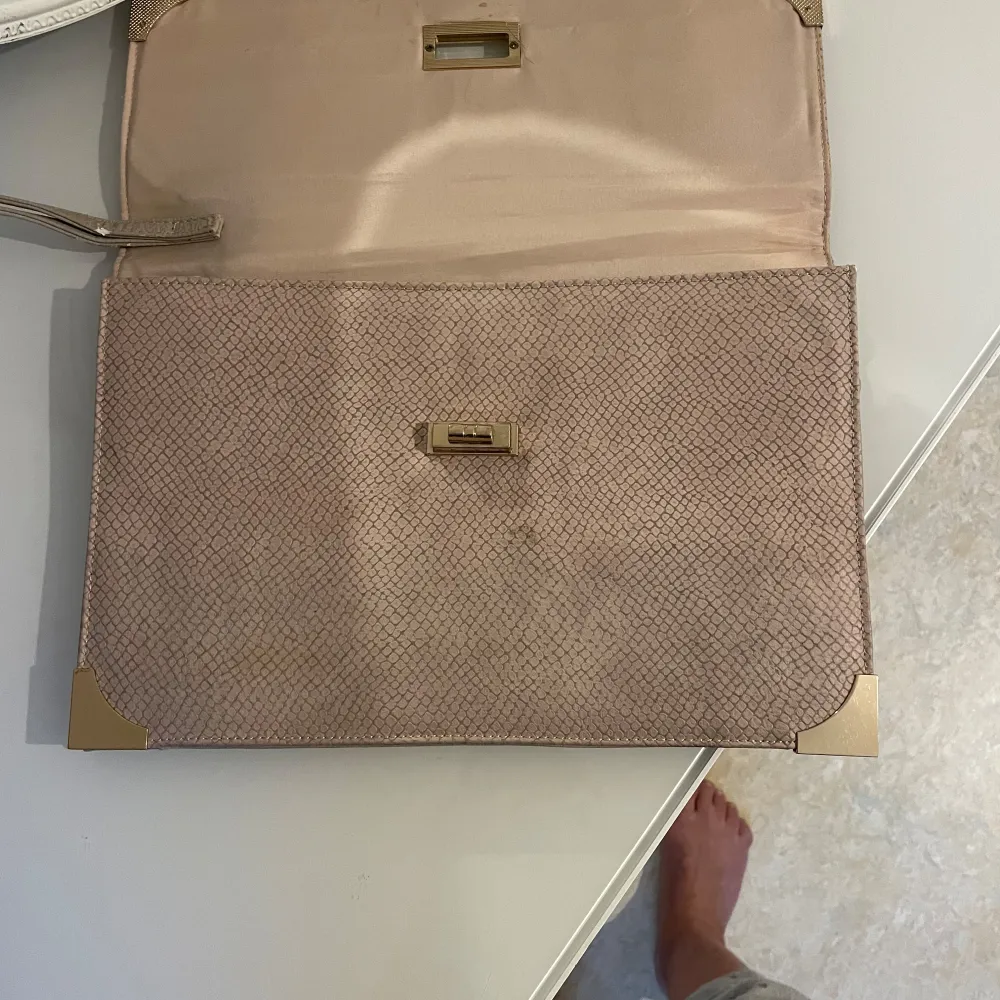 Elegant beige kuvertväska med guldfärgade hörndetaljer och ett stilrent lås framtill. Väskan har en diskret ormskinnsmönstrad yta och en praktisk handledsrem. Insidan är rymlig med fickor för småsaker. Perfekt för en chic look! Längd:22,22cm och bred : 33cm. Laukut & Käsilaukut.