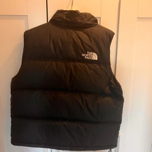 Svart dunväst från The North Face - Säljer en svart dunväst från The North Face. Västen har en hög krage och dragkedja framtill. Perfekt för kyligare dagar och ger en sportig look. Logotypen är synlig på bröstet och ryggen.