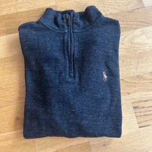 Polo Ralph Lauren half zip  - Säljer en stilren mörkgrå tröja från Ralph Lauren med dragkedja vid halsen. Tröjan har långa ärmar och en broderad logotyp på bröstet. Perfekt för en avslappnad och snygg look.