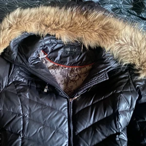 Svart dunjacka från Hollies - Snygg svart dunjacka från Hollies med pälskantad huva. Jackan har dragkedja och quiltad design, perfekt för kalla dagar. Den har praktiska fickor med knappar och dragkedjor. priset kan även diskuteras 😊😊