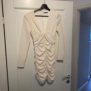 Beige klänning från Zara - Elegant beige klänning från Zara med långärmad design och v-ringad hals. Klänningen har en snygg rynkad detalj framtill som ger en unik look. Perfekt för en stilren och chic outfit.