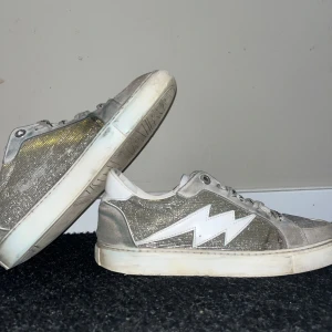 Glittriga sneakers från Zadig & Voltaire - Snygga sneakers med glittrig yta och en vit blixtdetalj på sidan. Skorna har snörning och en vit sula. Perfekta för att ge en cool touch till din outfit.