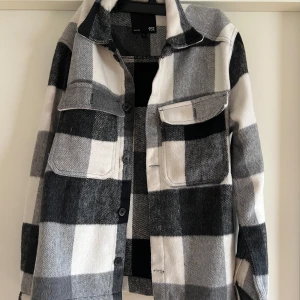 Rutig overshirt från lager 157 - Säljer en stilren rutig overshirt från lager 157 i svart, grått och vitt. Jackan har långa ärmar och knappar framtill samt två bröstfickor med knappar. Perfekt för lager-på-lager-stil.
