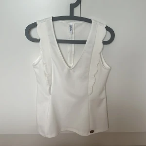 Vit ärmlös topp med dragkedja - Snygg vit ärmlös topp med v-ringning och dekorativa vågiga kanter. Toppen har en dragkedja i ryggen för enkel på- och avtagning. Perfekt för en stilren look.