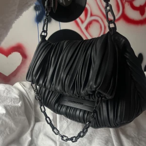 Svart axelväska från Karl Lagerfeld - Snygg svart axelväska från Karl Lagerfeld med ett coolt veckat mönster. Väskan har en kedja och en kort axelrem som går att justera, och en praktisk dragkedja på baksidan. Den köptes på Zalando för 2800kr och har bara använt några fåtal gånger🥳