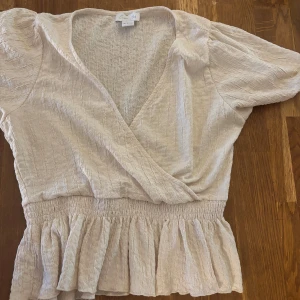 Vit  omlottopp med volangdetaljer - Säljer en söt vit omlottopp med korta ärmar och volangdetaljer i nederkant. Toppen har en v-ringad design och är tillverkad i ett lätt och luftigt material. Perfekt för varmare dagar eller för att klä upp sig lite extra.