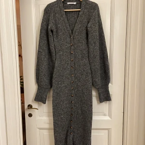 Grå  stickad klänning från ADOORE - Säljer en mysig grå lång stickad klänning från ADOORE med knappar hela vägen ner. Klänningen har långa ärmar och en v-ringad hals. Perfekt för kyligare dagar!