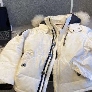 Vit dunjacka från Moose Knuckles - Säljer en snygg vit dunjacka från Moose Knuckles med pälsdetaljer på huvan. Jackan har dragkedja och knappar framtill samt broderad logga på insidan. Perfekt för kalla vinterdagar. Finns inget märken på axlen