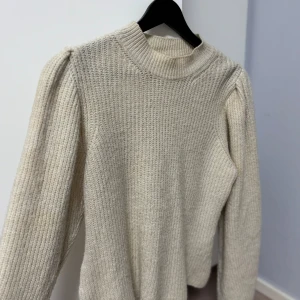 Beige stickad tröja från NLY Trend - Säljer en stilren beige stickad tröja från NLY Trend. Tröjan har en hög krage och långa ärmar med en lätt puffig design vid axlarna. Perfekt för kyligare dagar och ger en mysig känsla. Passar bra till både jeans och kjol.