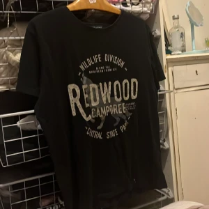 Svart t-shirt med tryck från Redwood - Säljer en svart t-shirt från Redwood med ett coolt tryck på framsidan. T-shirten har en rund hals och korta ärmar, perfekt för en avslappnad stil. Passar bra till jeans eller shorts!