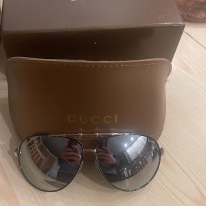 Gucci solglasögon med spegelglas - Snygga solglasögon från Gucci med spegelglas och klassisk aviatorform. De har en stilren design med en tunn metallram och kommer med ett brunt fodral med Gucci-logga. Perfekta för att ge en lyxig touch till din stil.