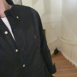 Svart skjorta från Polo Ralph Lauren - Snygg svart skjorta från Polo Ralph Lauren med klassisk krage och knappar framtill. Den har en broderad logotyp i rött på bröstet och är långärmad. Perfekt för en stilren look.