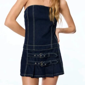 Axelbandslös jeansklänning med bälten - Trendig axelbandslös jeansklänning i mörkblå denim med två dekorativa bälten framtill. Perfekt för en cool och avslappnad stil. Klänningen har en kort längd och markerade sömmar som ger en snygg kontrast.