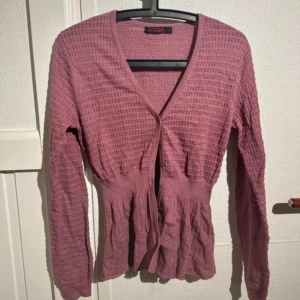 Rosa kofta från Indiska - Säljer en söt rosa kofta från Indiska med ett fint hålmönster. Koftan har långa ärmar och knappar framtill. Perfekt för att lägga till en färgklick i din garderob!