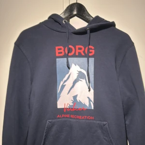 Mörkblå hoodie från Björn Borg - Säljer en mörkblå hoodie från Björn Borg med tryck av ett berg och texten 'Verbier Alpine Recreation' i rött. Tröjan har en klassisk känguruficka och justerbar huva med dragsko.