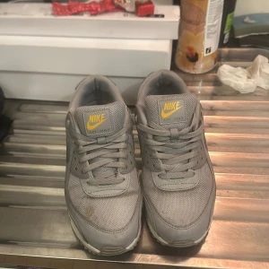 Grå Nike Air Max sneakers - Säljer ett par grå Nike Air Max sneakers med gula detaljer. Skorna har en klassisk design med snörning och en synlig luftkudde i sulan för extra komfort. Perfekta för en sportig look.