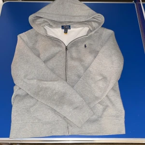 Grå hoodie från Polo Ralph Lauren - Säljer en snygg grå hoodie från Polo Ralph Lauren i storlek XL. Den är i mycket bra skick och har en dragkedja framtill. Perfekt för höst och vinter med sin mjuka och bekväma bomullskvalitet. Den klassiska loggan är broderad på bröstet. Passar både till vardags och chillkvällar. Är 175 och den passar perfekt.
