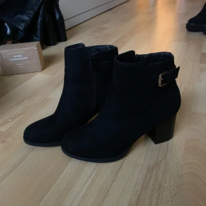 Svarta boots med klack och spänne - Snygga svarta boots i mocka med klack och dekorativt spänne i guld. De har en dragkedja på sidan. Perfekta för att ge en stilren touch till din outfit. Köpta på Din Sko för några år sedan och har använts 1 eller 2 gånger.
