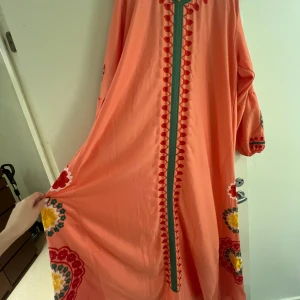 Helt ny aprikosrosa maxiklänning - Aldrig använd lång klänning /galabeya i aprikosrosa med broderier i rött, grönt och gult. Klänningen har långa ärmar och en rund halsringning. Den är löst sittande och har en bekväm passform.  Mjukt behagligt tyg 140 cm hals till fot Passar M/L/XL