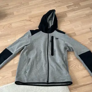 Säljer en snygg grå och svart hoodie från Nike. Den har en dragkedja framtill och en praktisk bröstficka med dragkedja. Perfekt för höst och vår, med en skön passform och huva för extra värme. Perfekt för både träning och vardag! Helt oanvändbar från Nike äkta!