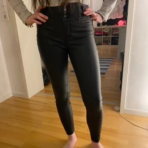 Byxor i läderimitation  - Säljer ett par svarta högmidjade skinny byxor i syntetmaterial. De har en snygg knapprad framtill och sitter tight för en smickrande passform. Perfekta för både vardag och fest! Det enda att anmärka är färgsläpp på knapparna. 