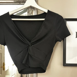 Svart kortärmad topp med knyt - Snygg svart topp med korta ärmar och en trendig knytning framtill. Perfekt för både vardag och fest! Den har en v-ringad design som ger en elegant touch. Passar bra till jeans eller kjol för en chic look.