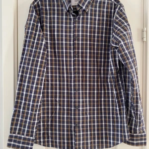 Ny grå/blå/orange rutig skjorta i strl 41 från The Shirt Factory, modell slim fit - Ny grå/blå/orange rutig skjorta i strl 41 från The Shirt Factory, modell slim fit.  Ny, aldrig använd.  Mått Över bröstet: ca 56 cm Längd från axeln och ner: ca 76 cm  100% bomull   Kommer från ett djur och rökfritt hem. 