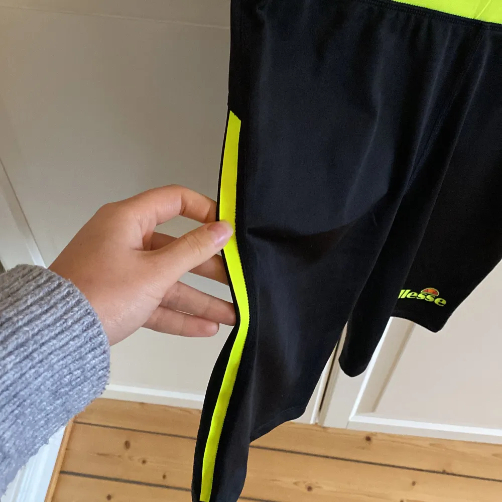 Säljer ett par svarta träningsshorts från Ellesse med en cool neongul linning och sidodetaljer. Perfekta för gymmet eller löprundan! De är bekväma och har en elastisk midja för bästa passform. Loggan syns tydligt på benet. 🏃‍♀️Storlek 38. I fint skick. . Sportswear.