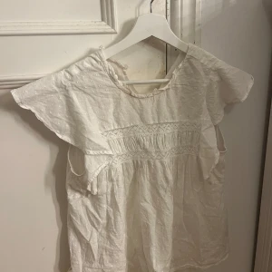 Vit blus med spetsdetaljer - Säljer en söt vit blus med spetsdetaljer och korta ärmar. Blusen har en lös passform och är gjord i ett lätt och luftigt material. Passar bra till både jeans och kjol!