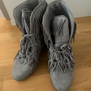 Grå mocka boots med fransar - Säljer ett par grå boots i mocka med snygga fransdetaljer. De har en inbyggd klack och snörning framtill. Perfekta för höst och vår! Använda men i bra skick! Storlek 40 men lite små i storleken❤️typ 38/39