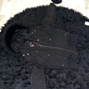 Svart pufferjacka från Moose Knuckles - Säljer en snygg svart pufferjacka från Moose Knuckles i storlek S. Jackan har en fluffig huva och är perfekt för kalla vinterdagar. Den har en dragkedja framtill och flera praktiska fickor. Perfekt för att hålla sig varm och stilren!