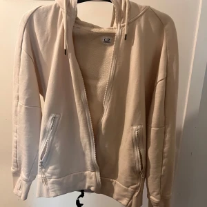 Beige hoodie från C.P. - Säljer en snygg beige hoodie från C.P. med dragkedja framtill och praktiska fickor med dragkedjor. Perfekt för en avslappnad stil och passar till alla årstider. Den har en skön passform och är supermjuk. Perfekt för både chill och aktivitet!