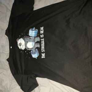Svart t-shirt med panda-tryck - Säljer en svart t-shirt i bomull med ett roligt tryck av en panda som lyfter vikter och texten 'The struggle is real'. Perfekt för gymmet eller som en avslappnad vardagströja. T-shirten är i bra skick och har korta ärmar.