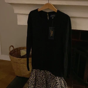 Svart klänning med blommigt mönster från Polo Ralph Lauren - Snygg svart klänning från Polo Ralph Lauren med ett blommigt mönster i nederdelen. Klänningen har långa ärmar och är i nytt skick med etikett kvar. Obs det är en barnklänning och passar 8-10 åringar. 