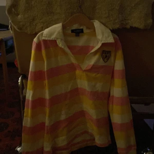 Randig pikétröja från GANT - Säljer en färgglad randig pikétröja från GANT i storlek XL för 11-12 år. Tröjan har breda ränder i rosa, gul och vit med en klassisk krage och knappar framtill. Perfekt för vår och sommar! 🏷️