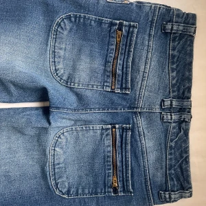 Blå jeans med dragkedjor och knappar - Säljer ett par snygga lågmidjade blå jeans i storlek 36. De har coola dragkedjor på bakfickorna och dubbla knappar fram. Perfekta för en avslappnad stil med en touch av edge. Passar bra till både vardag och fest!